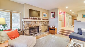 A living room with a stone fireplace, a sofa, a coffee table, and a television mounted on the wall. - Lake Tahoe Corner Condo w/ Hot Tub, Beaches Nearby (Incline Village)
