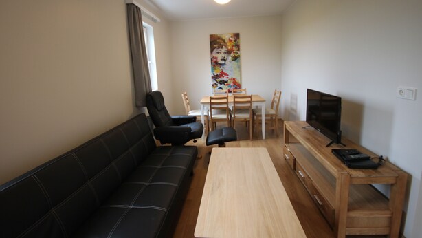 A living room with a black leather sofa, a wooden coffee table, a TV on a wooden stand, and a dining area with a table and chairs. - Selfell Guesthouse - Apartment 2 (Kirkjubæjarklaustur)