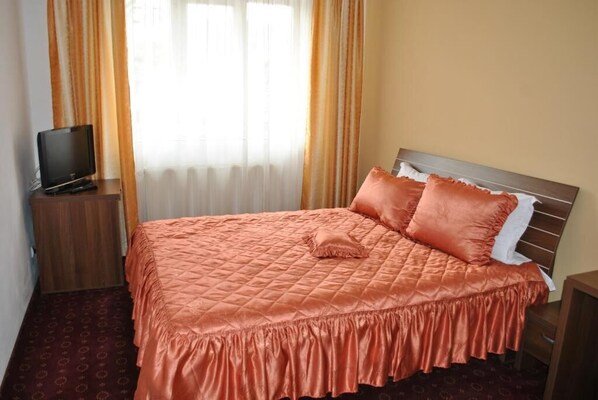 A neatly made bed with a quilted cover, a television on a wooden nightstand, and a window with curtains.