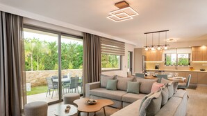Un salon moderne comprenant un canapé, un pouf et une table basse, un coin repas avec des chaises, et une cuisine avec des meubles en bois et des luminaires suspendus.