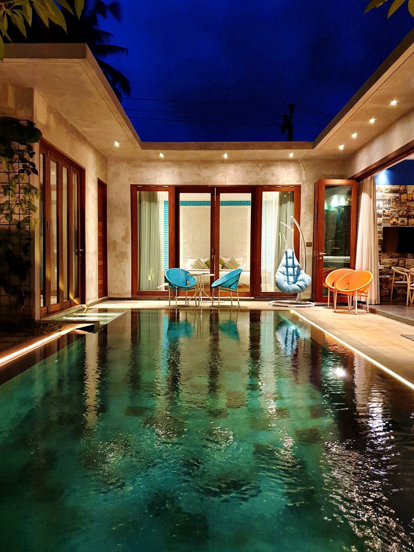 Una piscina interior con agua cristalina, rodeada de puertas de madera y paneles de vidrio, y amueblada con sillas azules y anaranjadas.