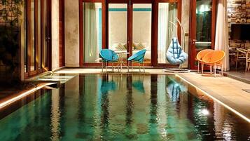 Una piscina interior con agua cristalina, rodeada de puertas de madera y paneles de vidrio, y amueblada con sillas azules y anaranjadas.