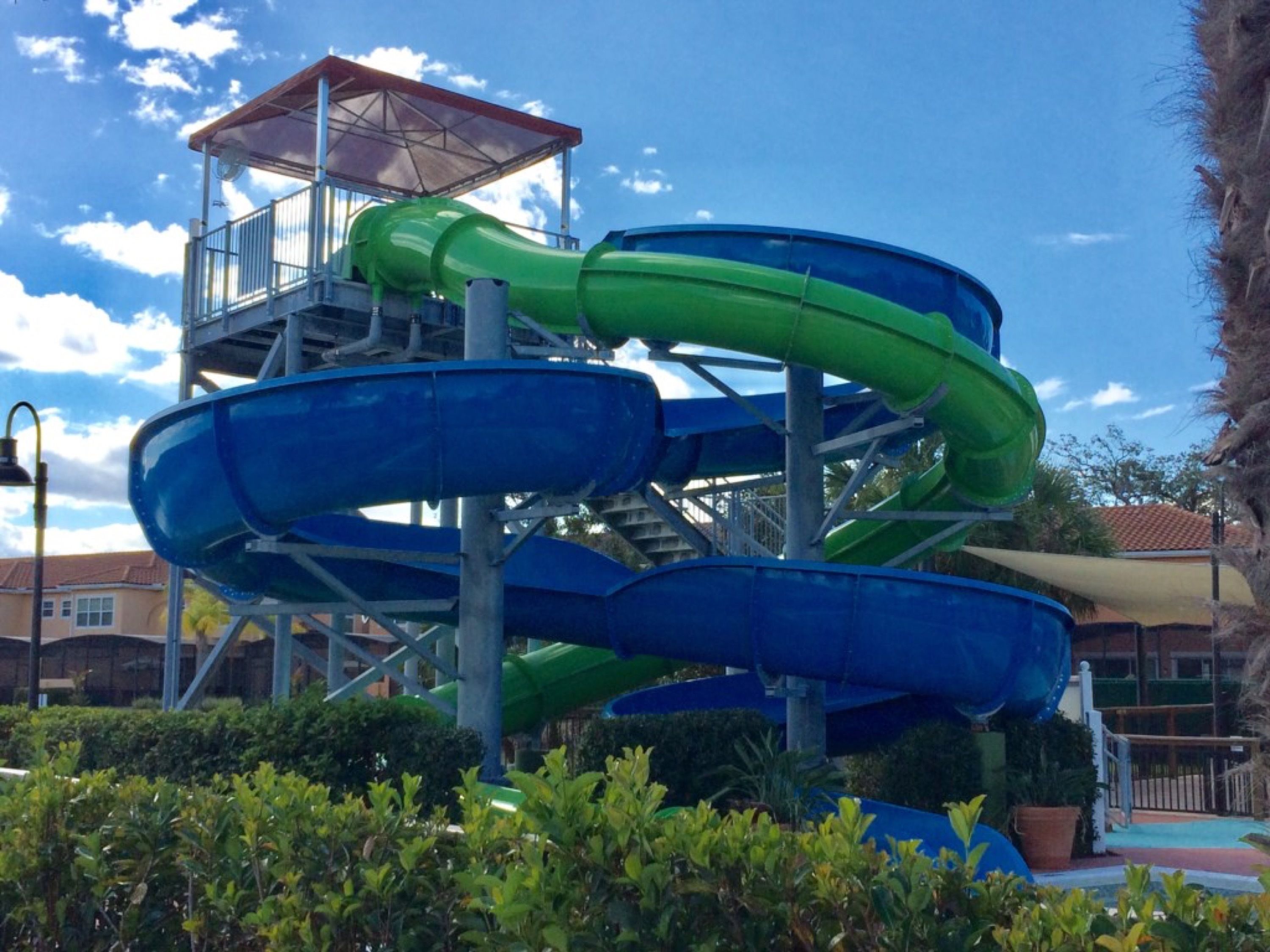 A multi-level water slide structure with green and blue slides, surrounded by greenery and a clear sky.