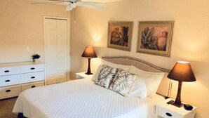 Une chambre à coucher comprenant un lit, deux tables de chevet, un ventilateur de plafond et deux tableaux accrochés au mur.