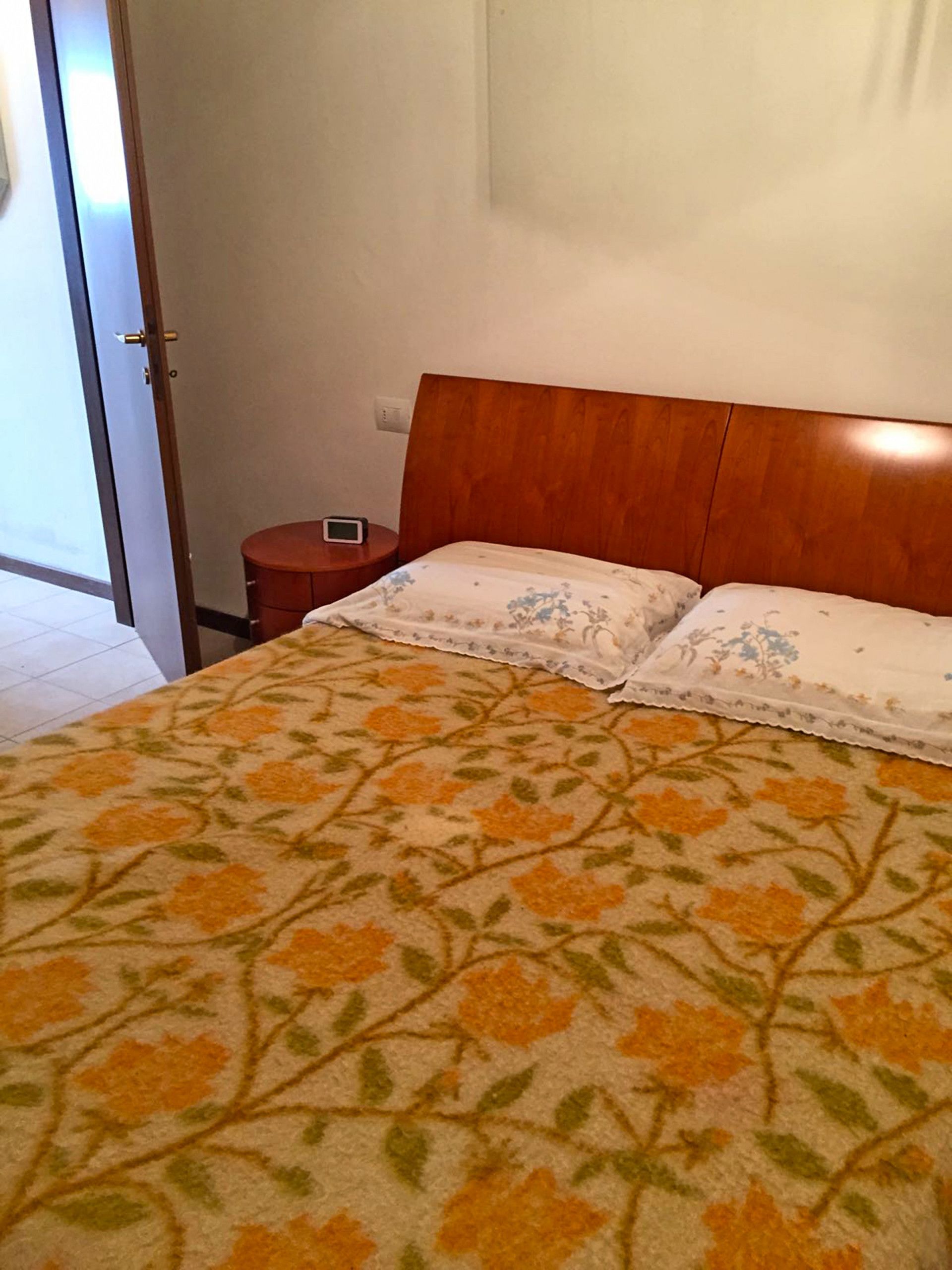 A bedroom with a wooden headboard, a bed with floral-patterned bedding, a bedside table with a clock, and a door leading to another room.