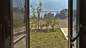 A view from an open door to a balcony with a small garden, a distant building, and a mountain in the background. - La Quiete 30 Holideal (Tremosine)