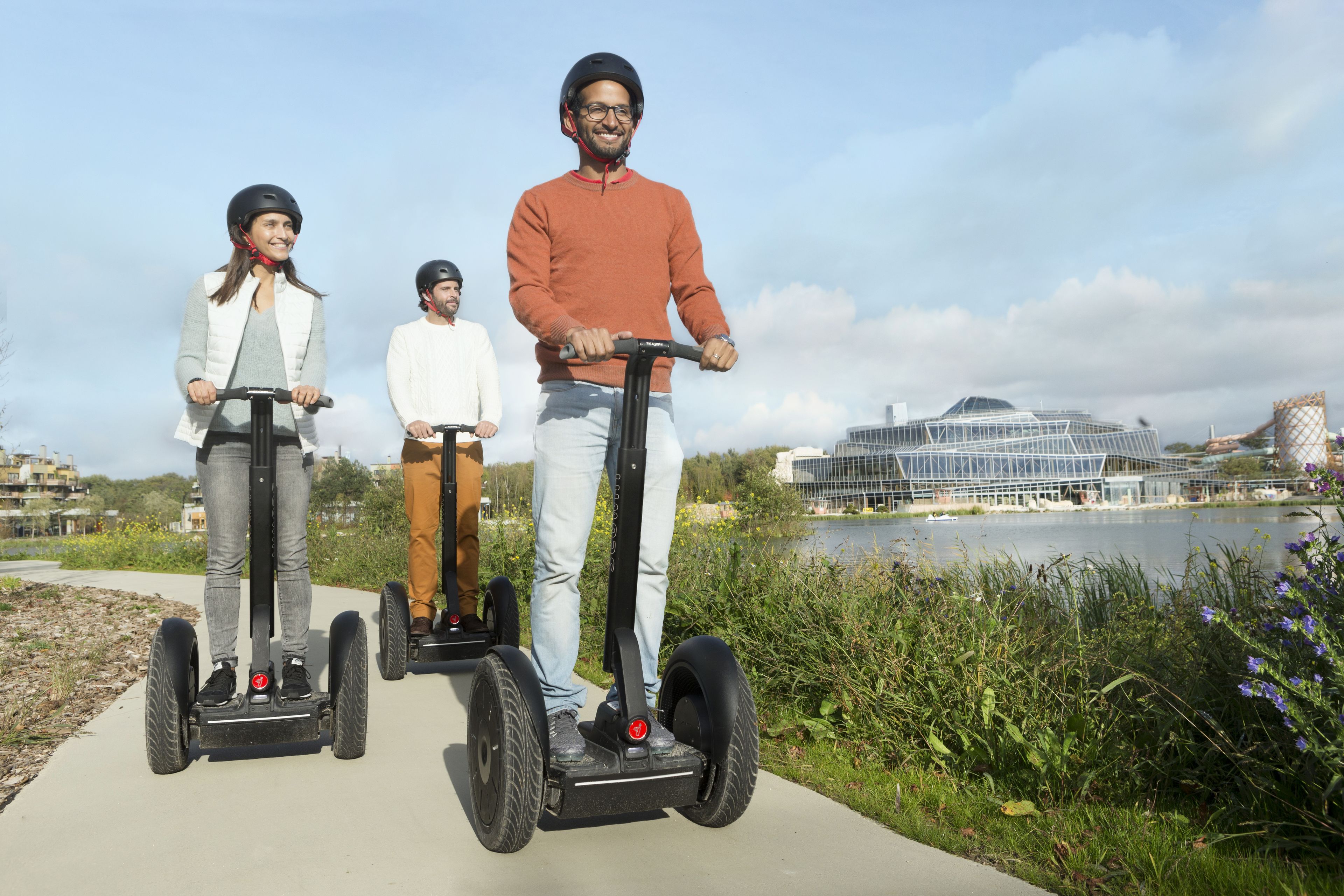 Tres personas montando segways por un sendero, con un edificio moderno y una masa de agua al fondo.