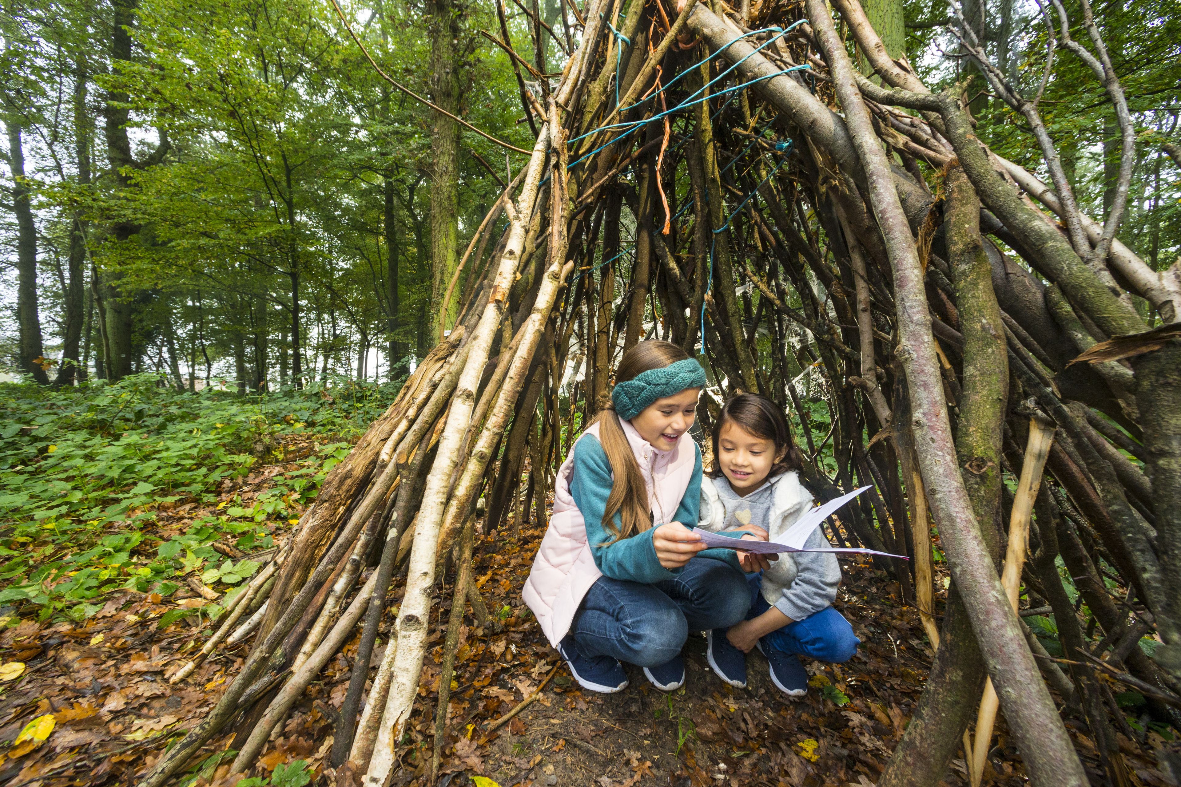 Two people sitting under a natural structure made of branches in a forest.