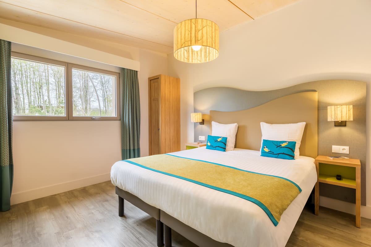 A hotel room with a large bed, two bedside lamps, a nightstand, a window with curtains, and a ceiling light fixture.