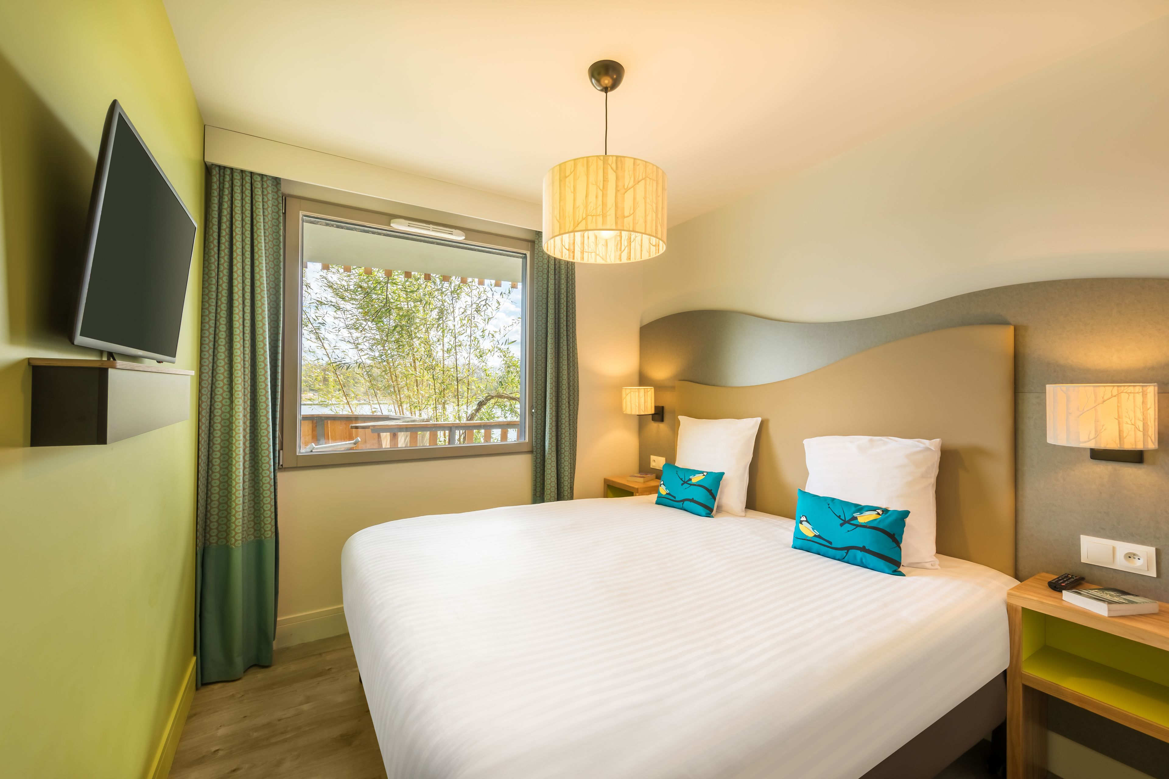 A hotel room with a large bed, a television on a stand, a window with curtains, a lamp, and a nightstand with a book and remote control.