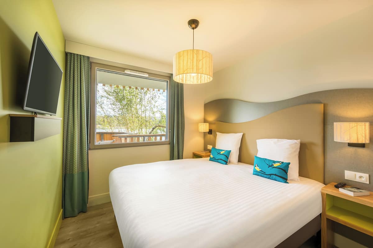 A hotel room with a large bed, a television on the wall, a window with curtains, and a bedside table with a lamp.