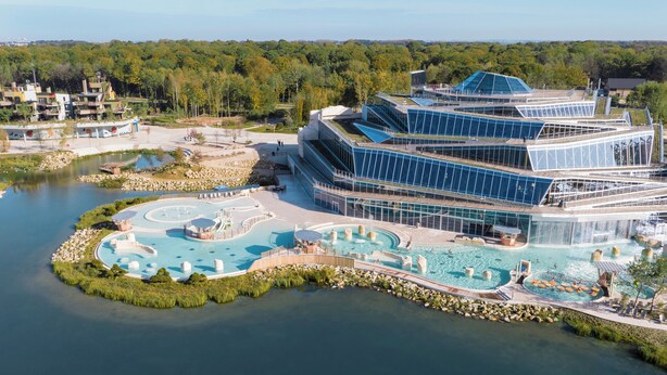 A modern hotel with a waterfront pool area, including a circular pool and a larger central pool, surrounded by lush greenery and a clear sky. - Les Villages Nature Paris (Villeneuve-le-Comte)