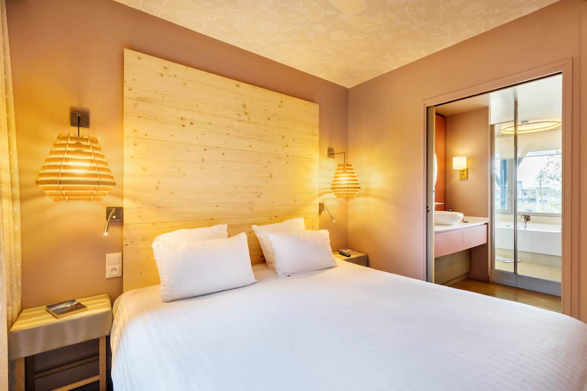 A modern hotel room with a large bed, wooden headboard, bedside lamps, a nightstand, and a bathroom with a glass door.