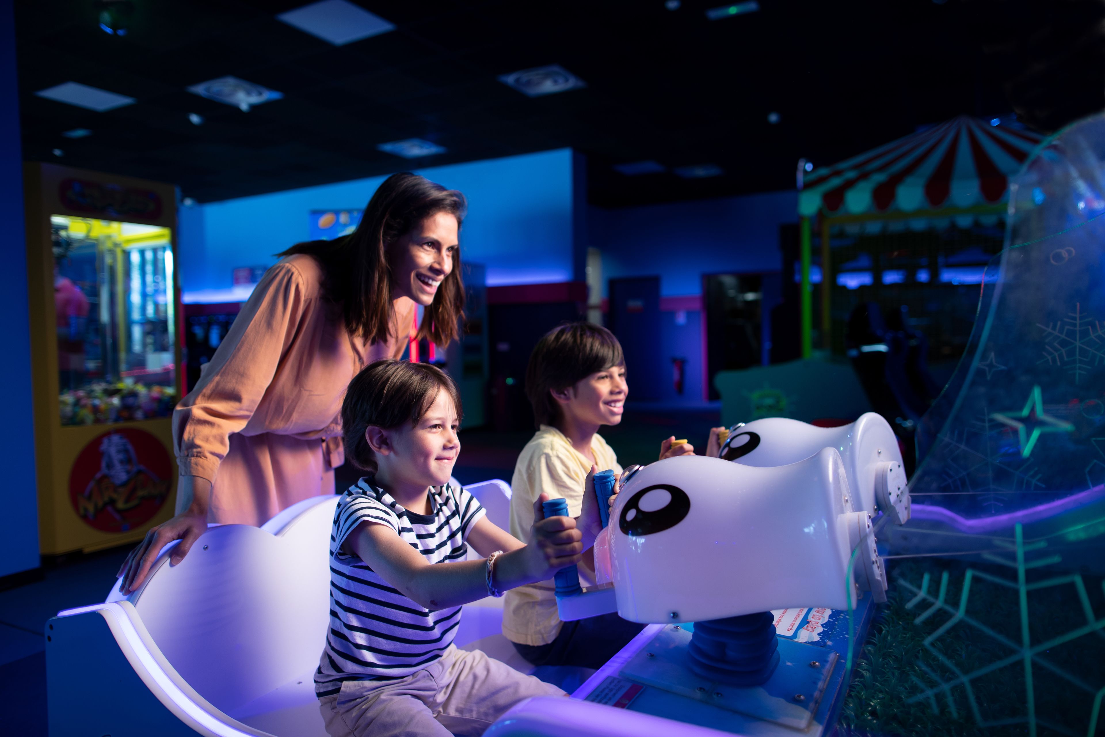 Una famiglia gioca a un videogioco in un'area giochi indoor, con cabinati arcade e una macchina a grinfie sullo sfondo.