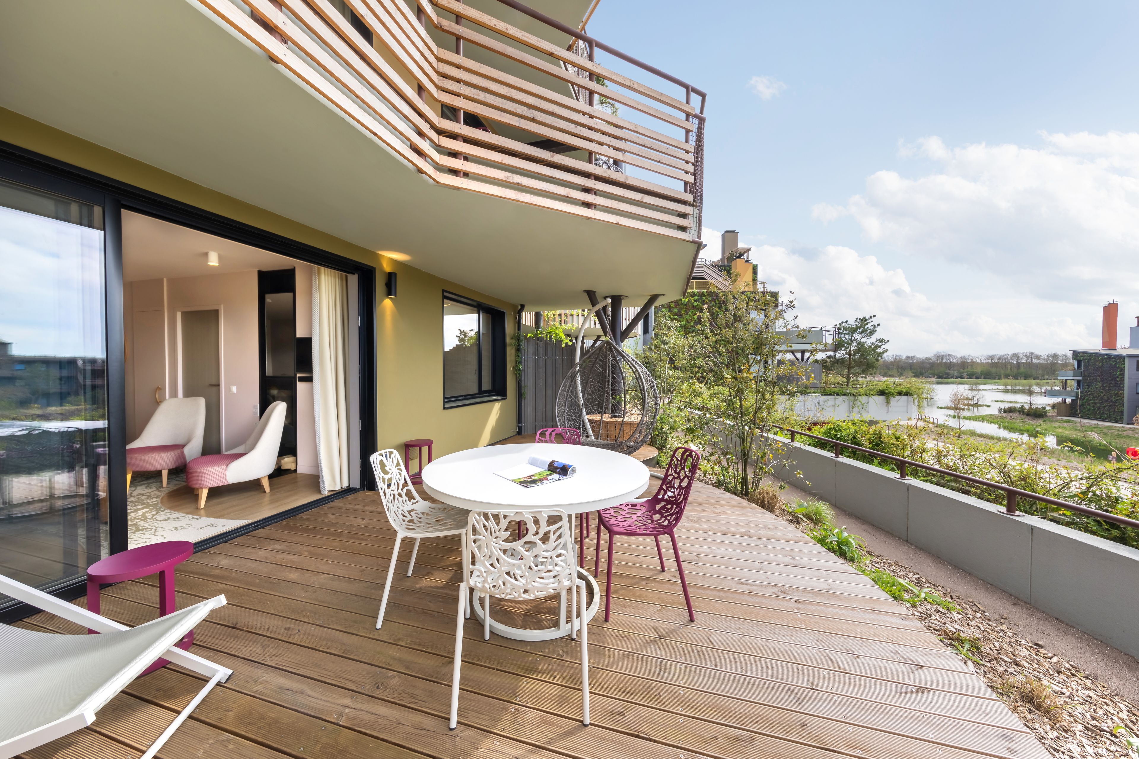 A modern outdoor deck with a round table and chairs, a glass door leading to an interior space, and a view of a water body.