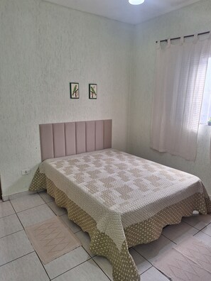Ein Schlafzimmer mit einem Bett, einem Kopfteil, einem Fenster mit Vorhängen und zwei an der Wand befestigten Bildern.