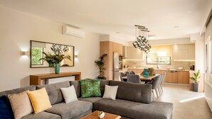 A modern living room with a grey sectional sofa, wooden coffee table, and a dining area with a table and chairs. A kitchen with wooden cabinets and a refrigerator is visible in the background. - Kagiemar Luxury House (Chania)