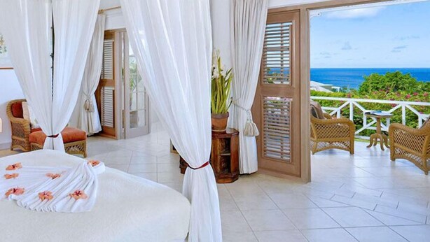 Une chambre d’hôtel avec un lit, un balcon meublé de mobilier en osier et une vue sur l’océan.