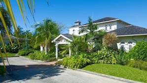 A large, two-story house with a white exterior and dark roof, surrounded by lush greenery and palm trees. - Villa Oceana (Holetown)