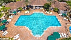 Uma piscina retangular grande cercada por cadeiras de descanso e guarda-sóis, com um pavilhão coberto no centro e uma área residencial ao fundo.