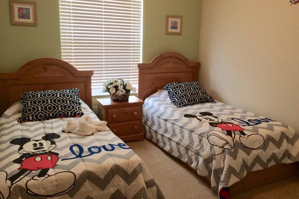 Two twin beds with Mickey Mouse bedding, a wooden nightstand with a vase of flowers, and a window with blinds.