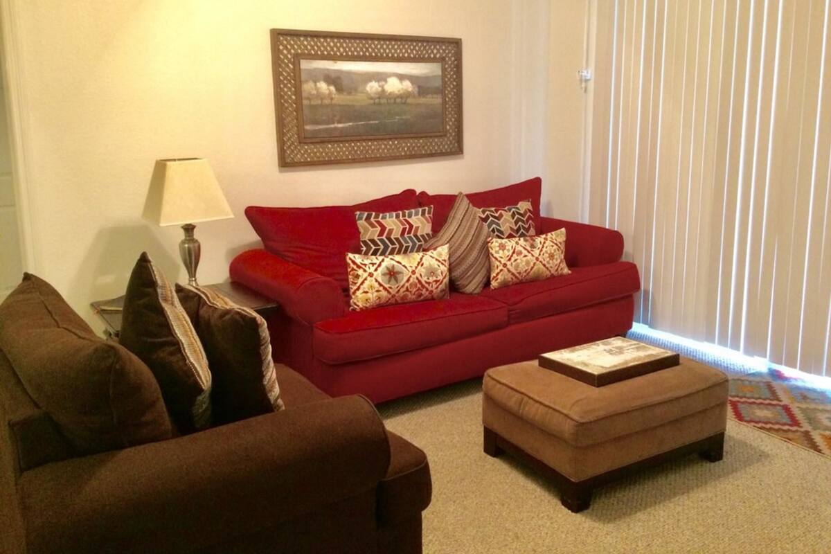 A living room with a red sofa, brown armchair, and ottoman. There is a lamp, a framed picture on the wall, and a window with blinds.
