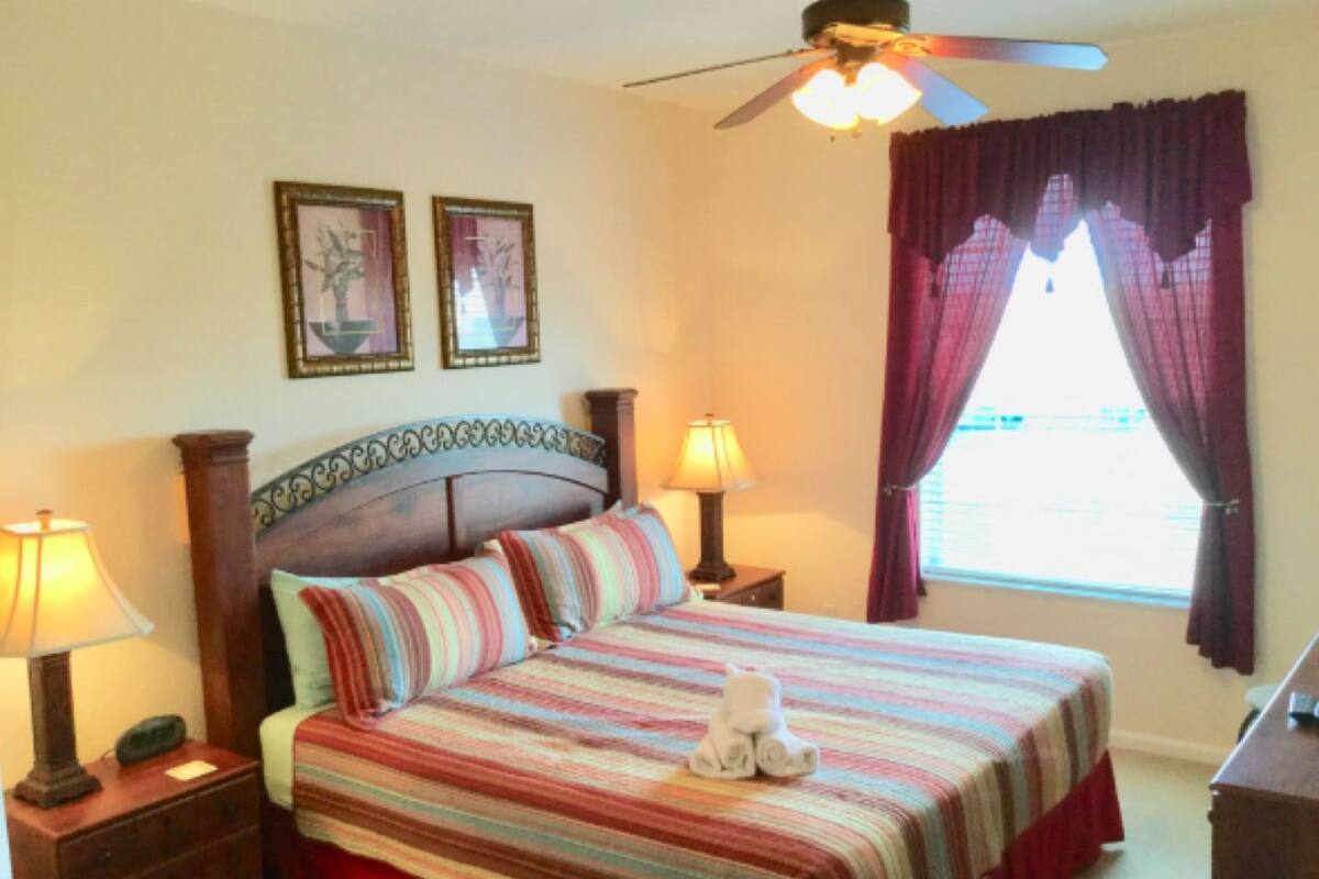 A bedroom with a bed, two bedside lamps, a ceiling fan, and a window with curtains.
