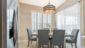 Uma sala de jantar com uma mesa de madeira, quatro cadeiras cinzas, um lustre pendente e uma janela grande com persianas.