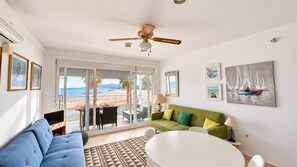Una sala de estar con sofá, un diván verde, una mesa de centro y una televisión. Cuenta con un balcón con vistas a la playa y palmeras.