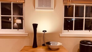 Una mesa de madera con un jarrón negro y un cuenco blanco, ubicada entre dos ventanas con marcos blancos y un pequeño cuadro encima.