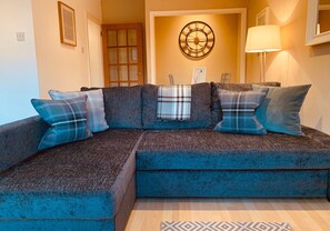 A sectional sofa with blue and plaid cushions, a wall clock, and a floor lamp in a room with a wooden floor and a patterned rug.