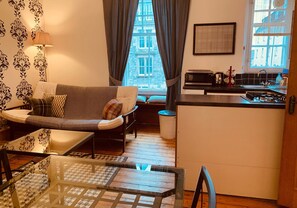 Un espace de vie compact comprenant un canapé, une table basse en verre, un coin cuisine avec un four à micro-ondes et une vue sur un immeuble à travers de grandes fenêtres.
