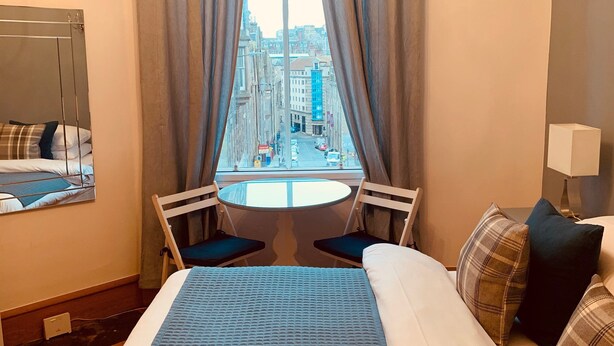 A hotel room with a bed, a small round table, two chairs, a window with a city view, and a lamp.