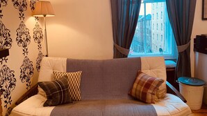 A cozy living room with a sofa, patterned wallpaper, a lamp, and a view of buildings through a window.