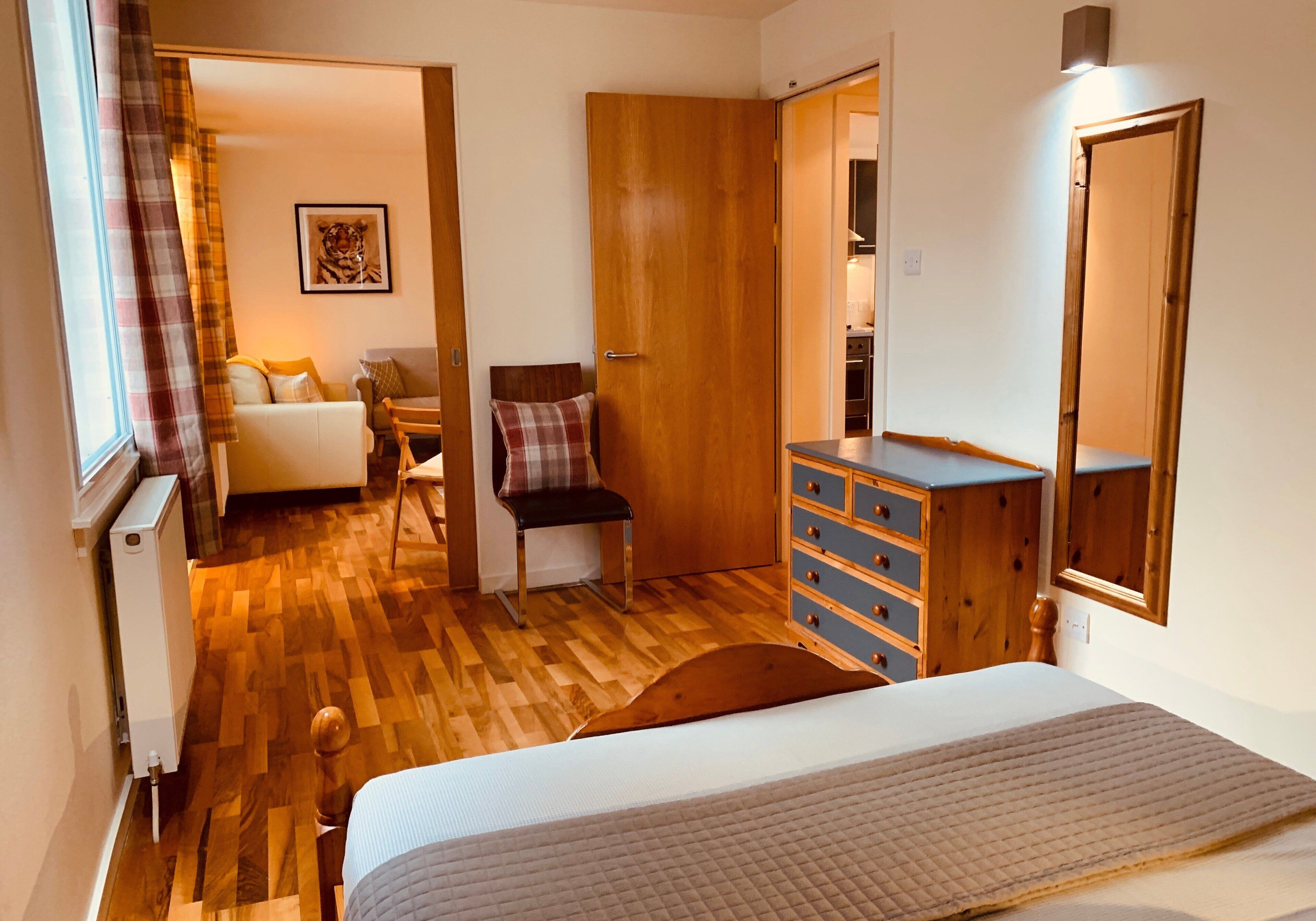 Un dormitorio con cama, cómoda de madera y una silla. Se ve una puerta que da a otra habitación, un radiador y un cuadro en la pared.
