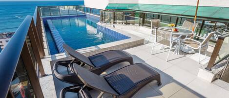 A rooftop pool area with lounge chairs, a table with fruit, and a view of the ocean.