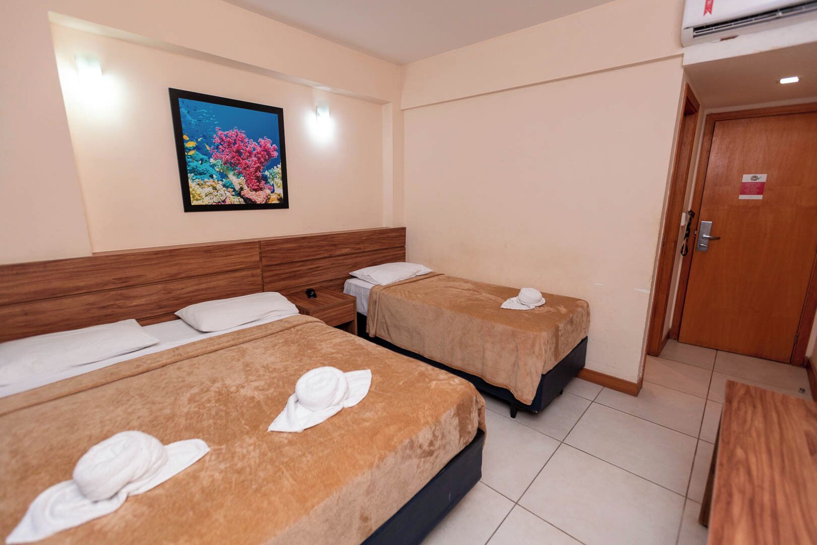 A hotel room with two beds, a wooden headboard, a painting of an underwater scene, and a door with a sign.