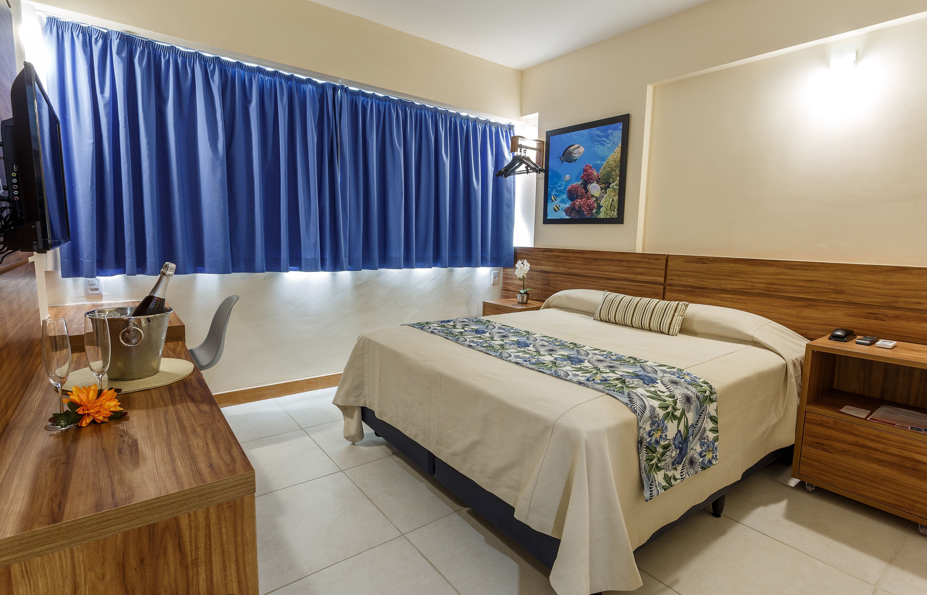 A hotel room with a bed, a wooden desk, a chair, a nightstand, and a window with blue curtains.