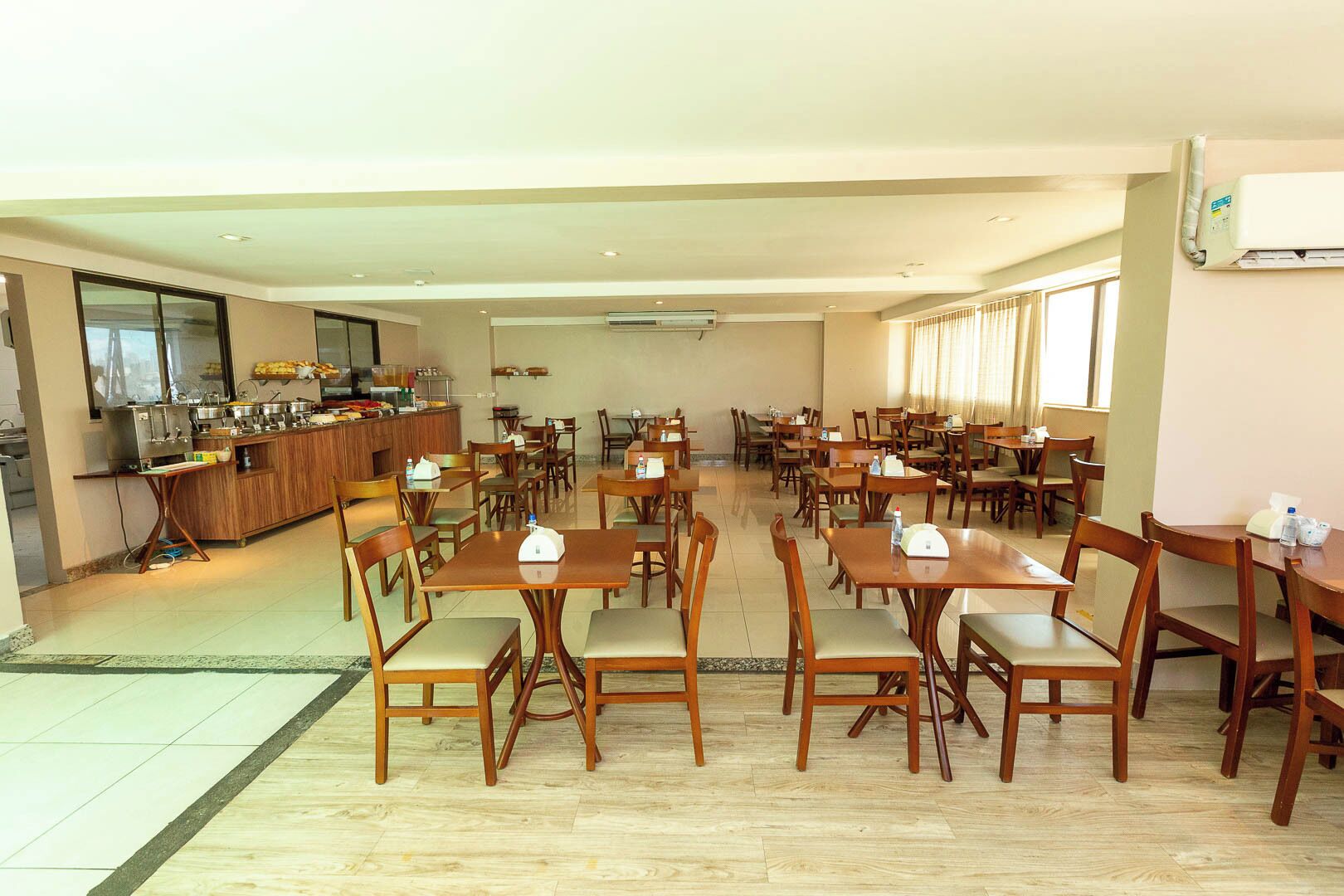 Un comedor amplio con varias mesas y sillas, una zona de cocina con bufé, y un ventanal grande que proporciona luz natural.