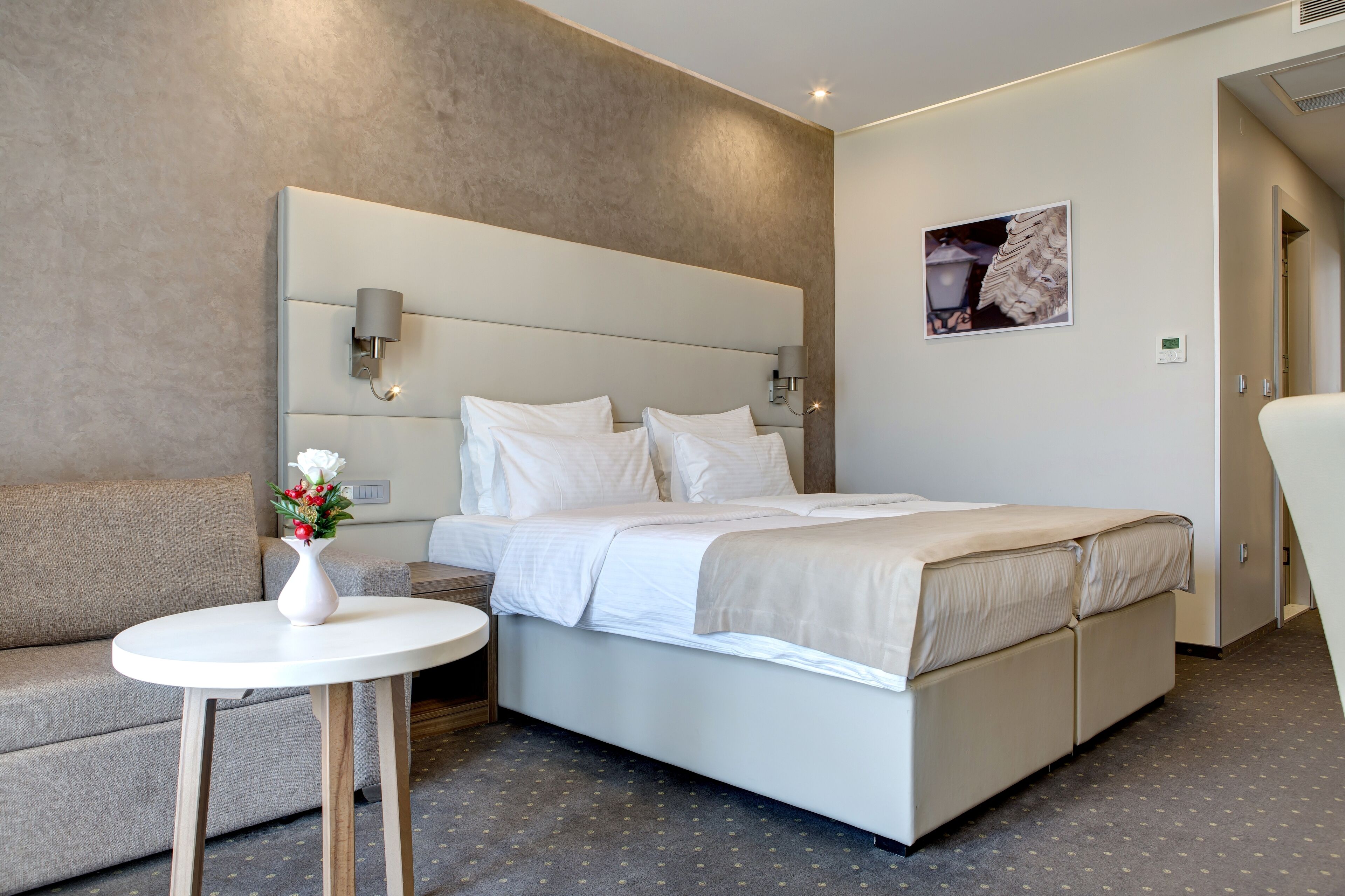 Quarto de hotel moderno com uma cama grande, uma mesa redonda pequena, um sofá e um quadro na parede.