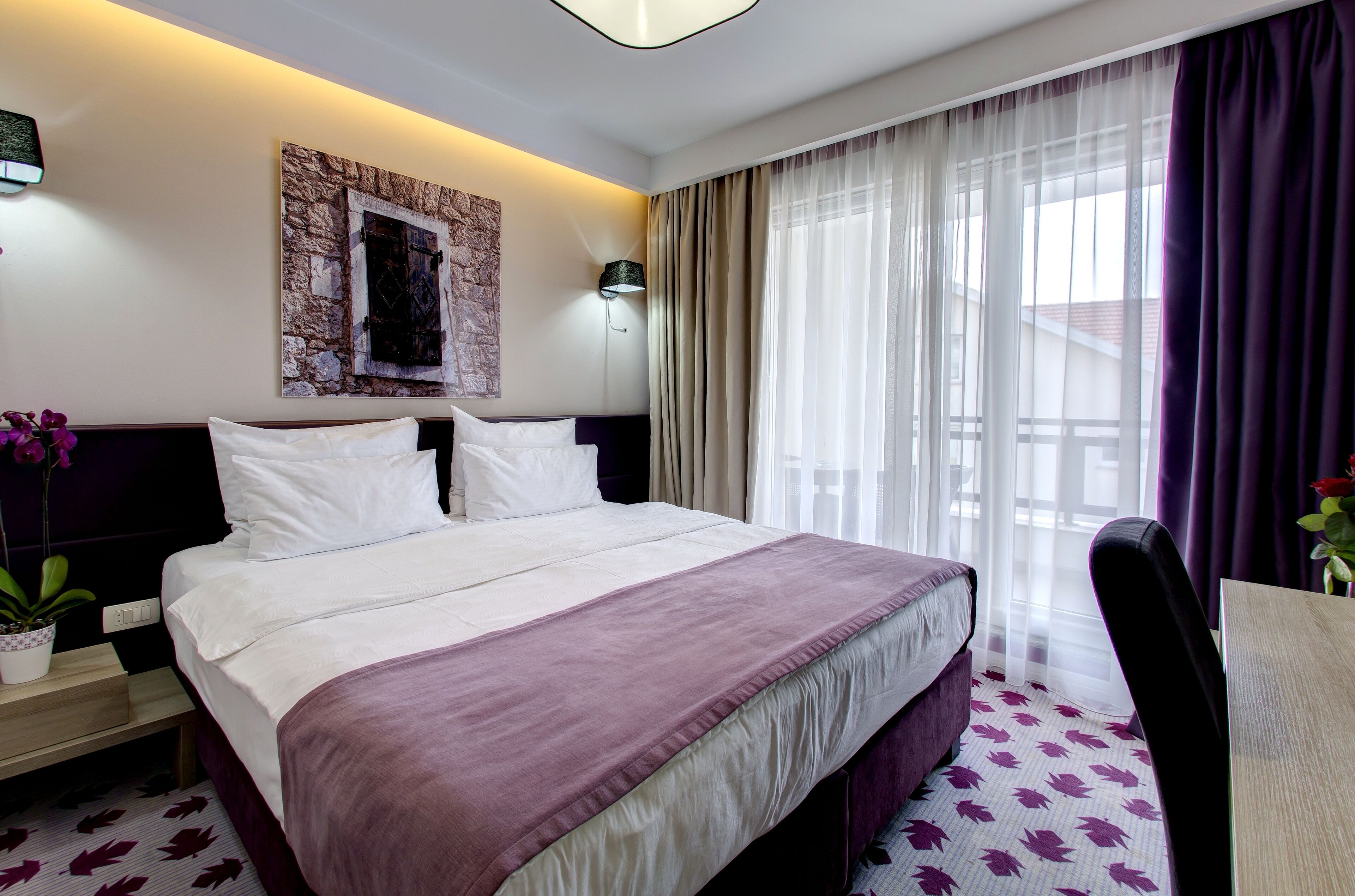 A hotel room with a large bed, a nightstand, a lamp, a window with sheer curtains, and a view of the outside.