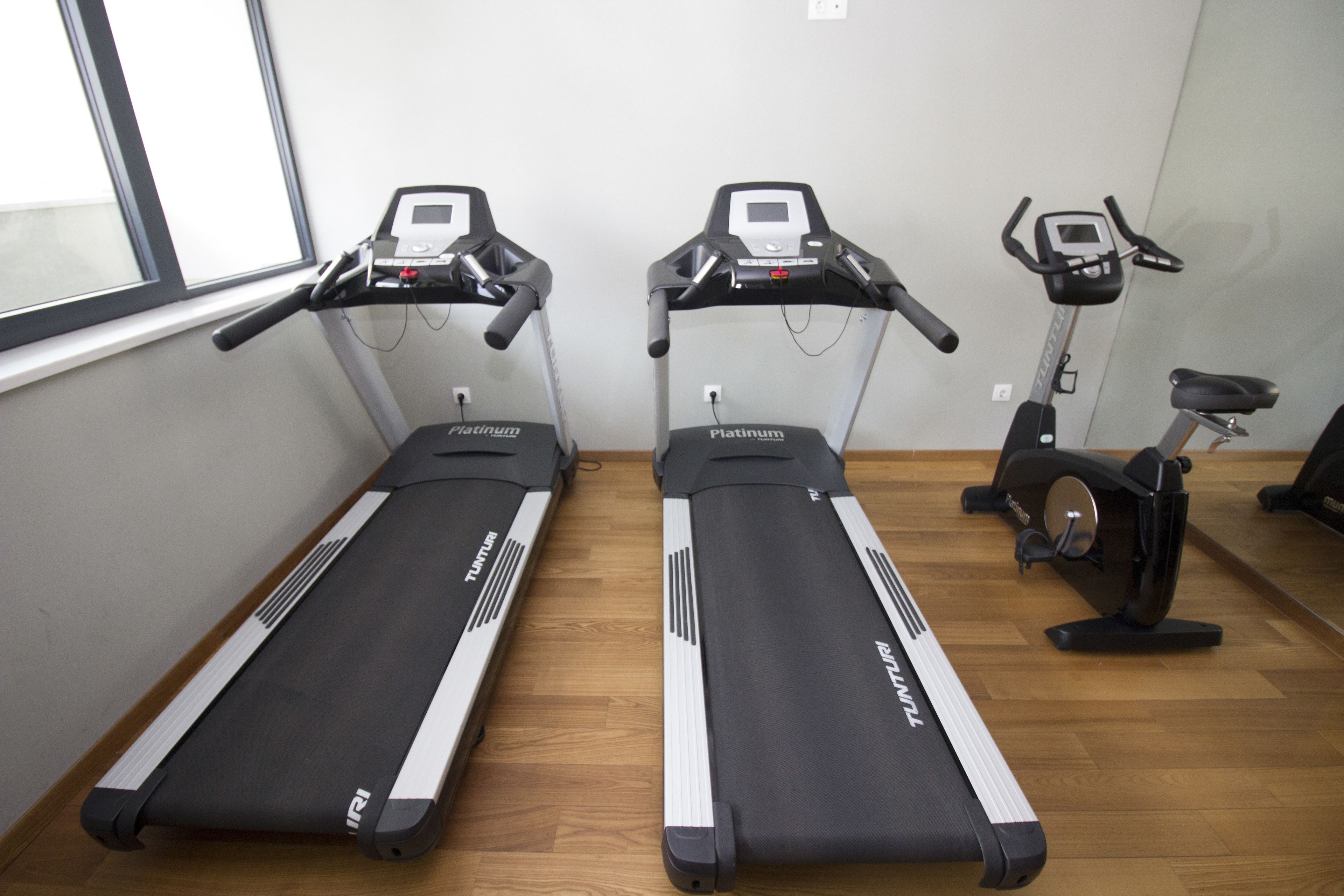 Two treadmills and an elliptical machine in a room with wooden flooring and a window.