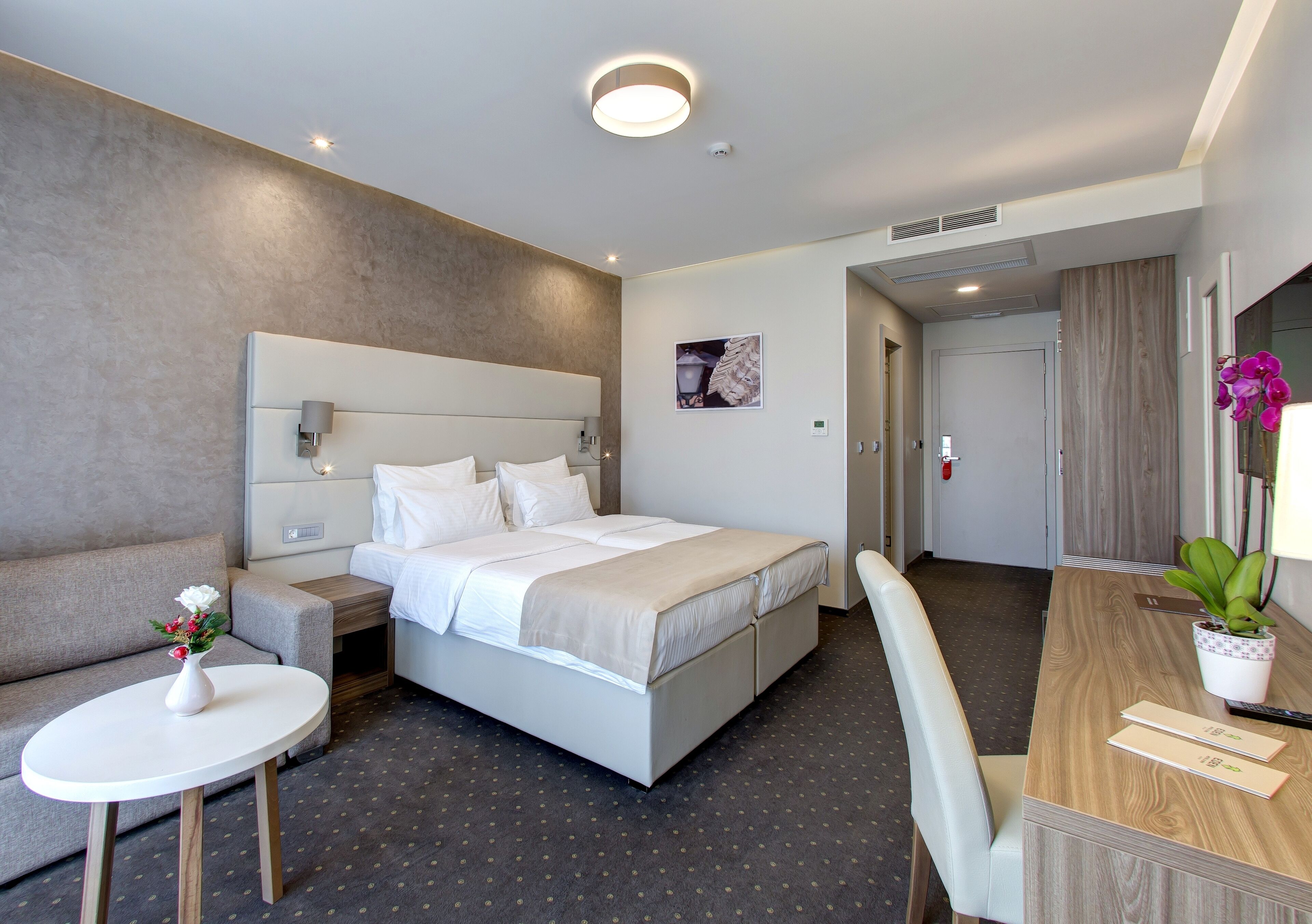 A modern hotel room with a bed, a small table with a vase of flowers, a desk with a plant, and a couch.