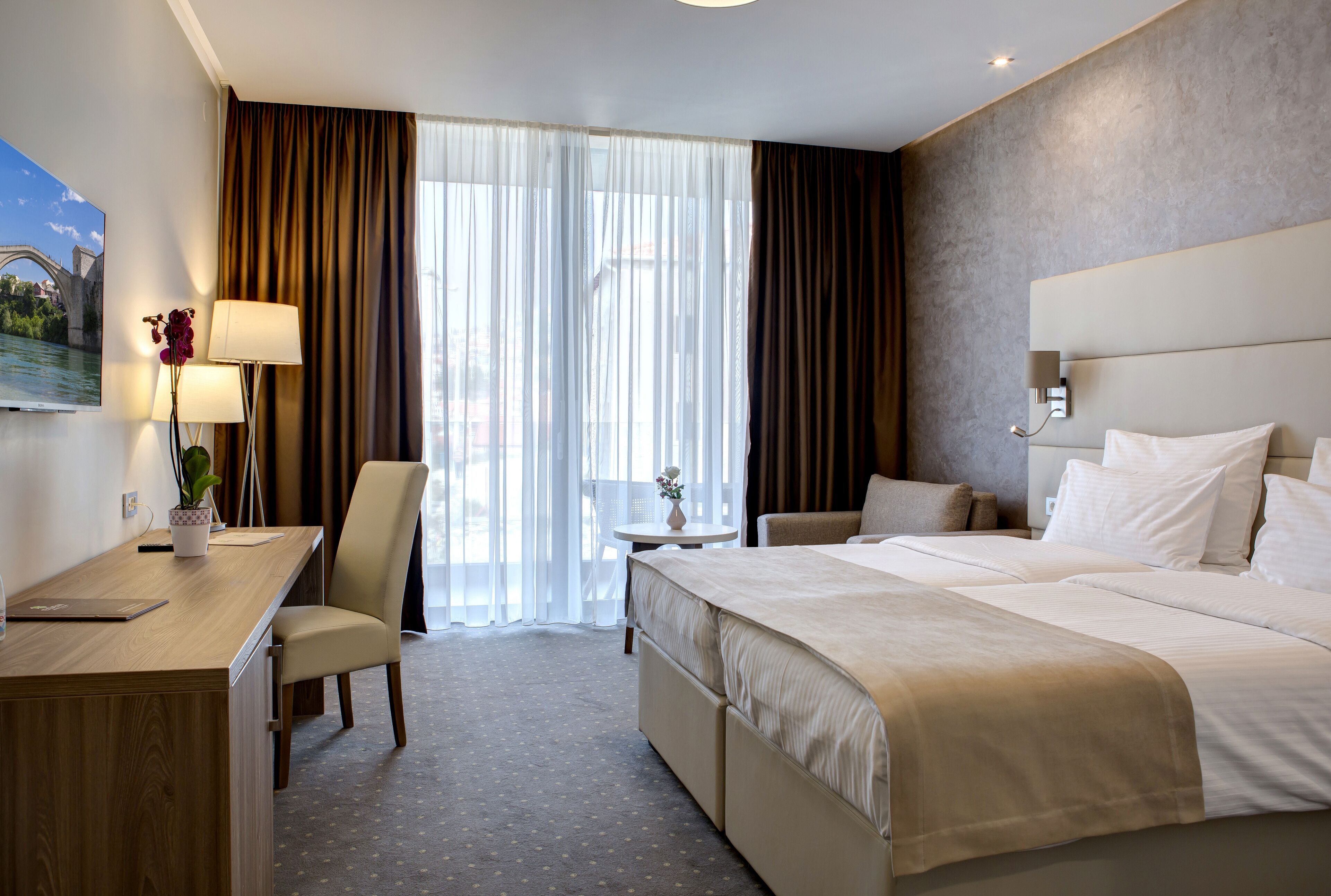 Quarto de hotel com uma cama grande, uma escrivaninha com um laptop, uma cadeira, uma pequena mesa com um vaso e vista para uma ponte pela janela.