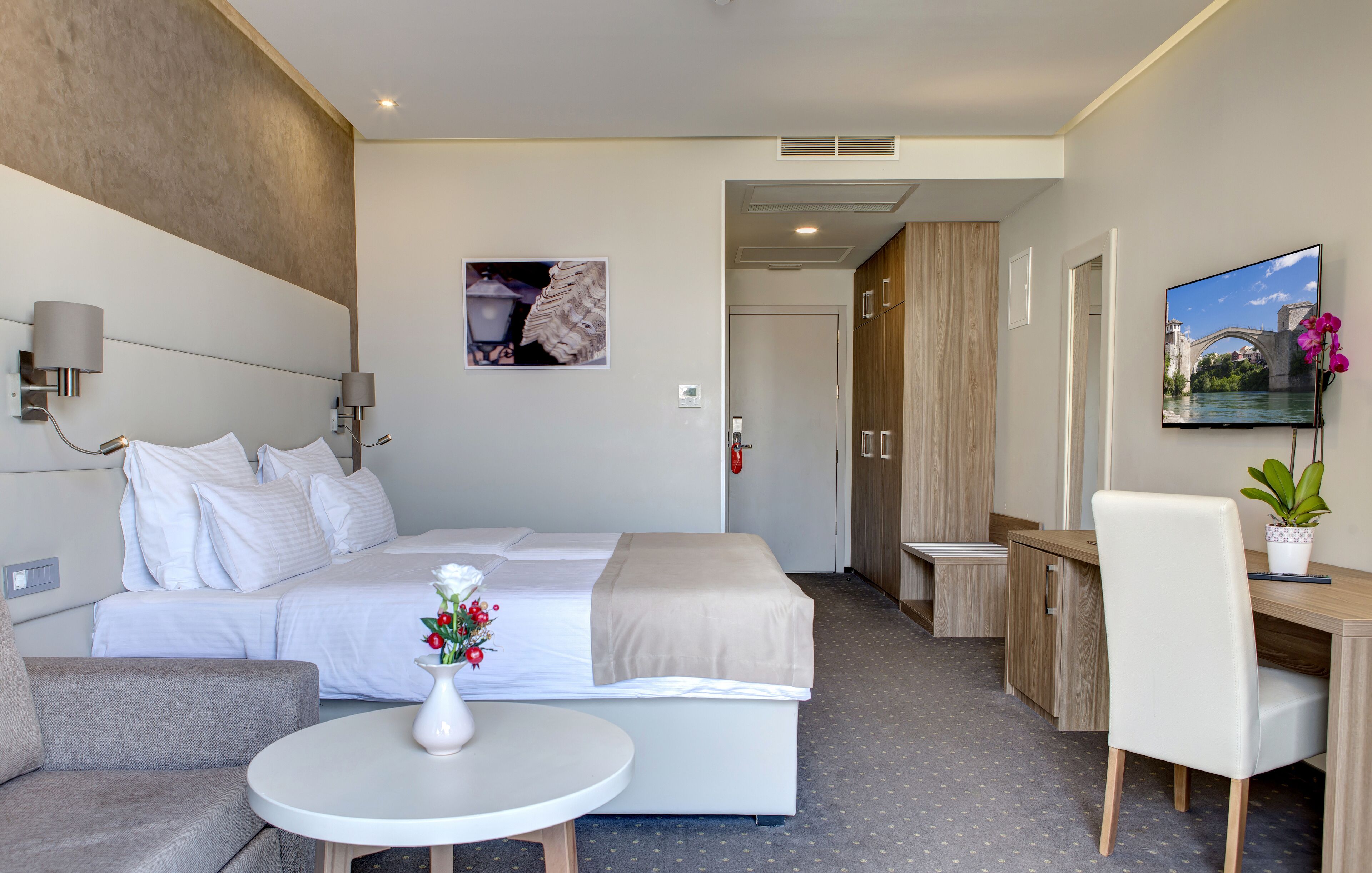 Habitación de hotel con una cama, un escritorio, una silla, una mesita con un florero, un televisor y un cuadro en la pared.