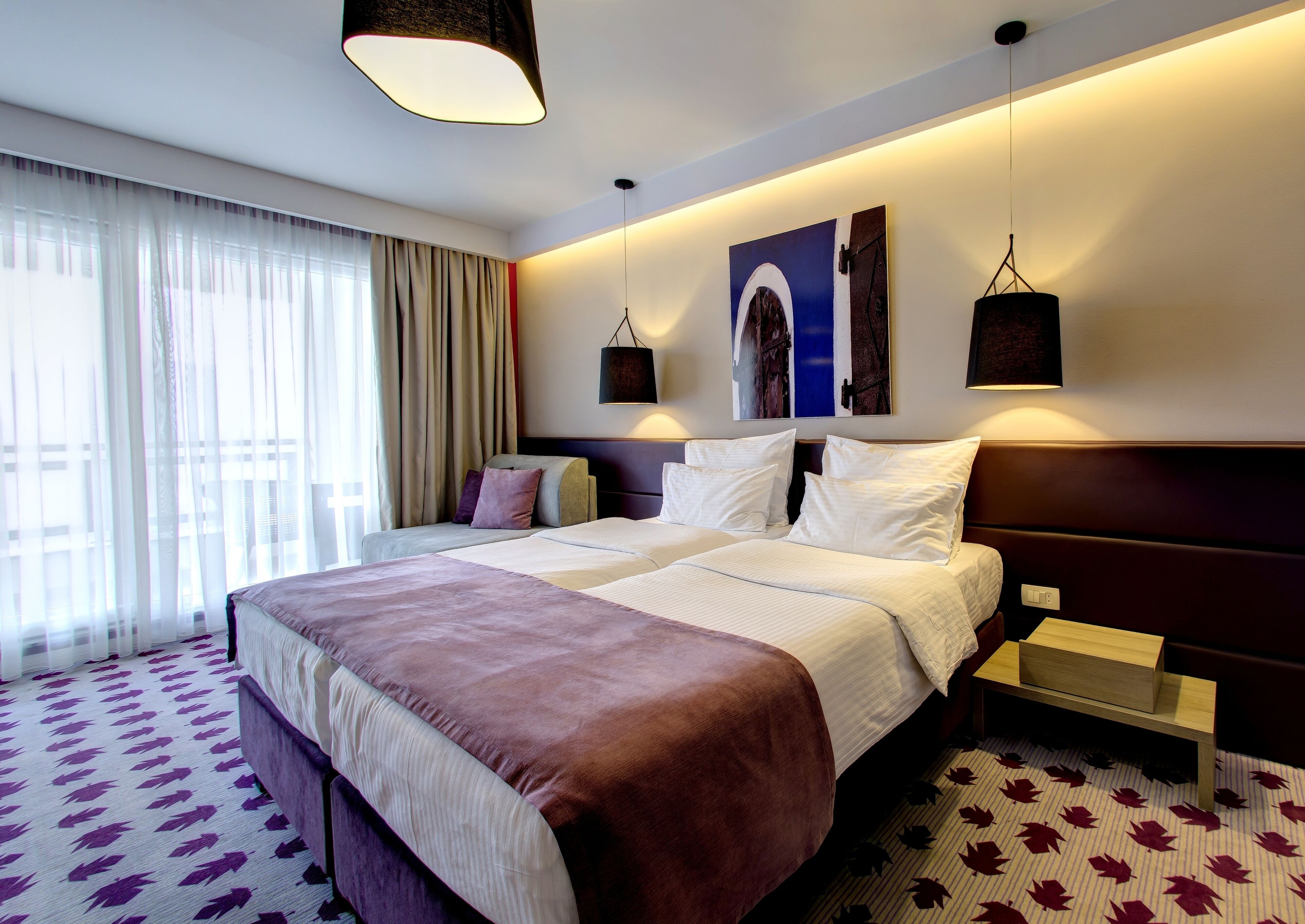 Habitació d'hotel amb un llit gran, dues taules de nit, una tauleta auxiliar, una finestra amb cortines transparents i una obra d'art penjada a la paret.