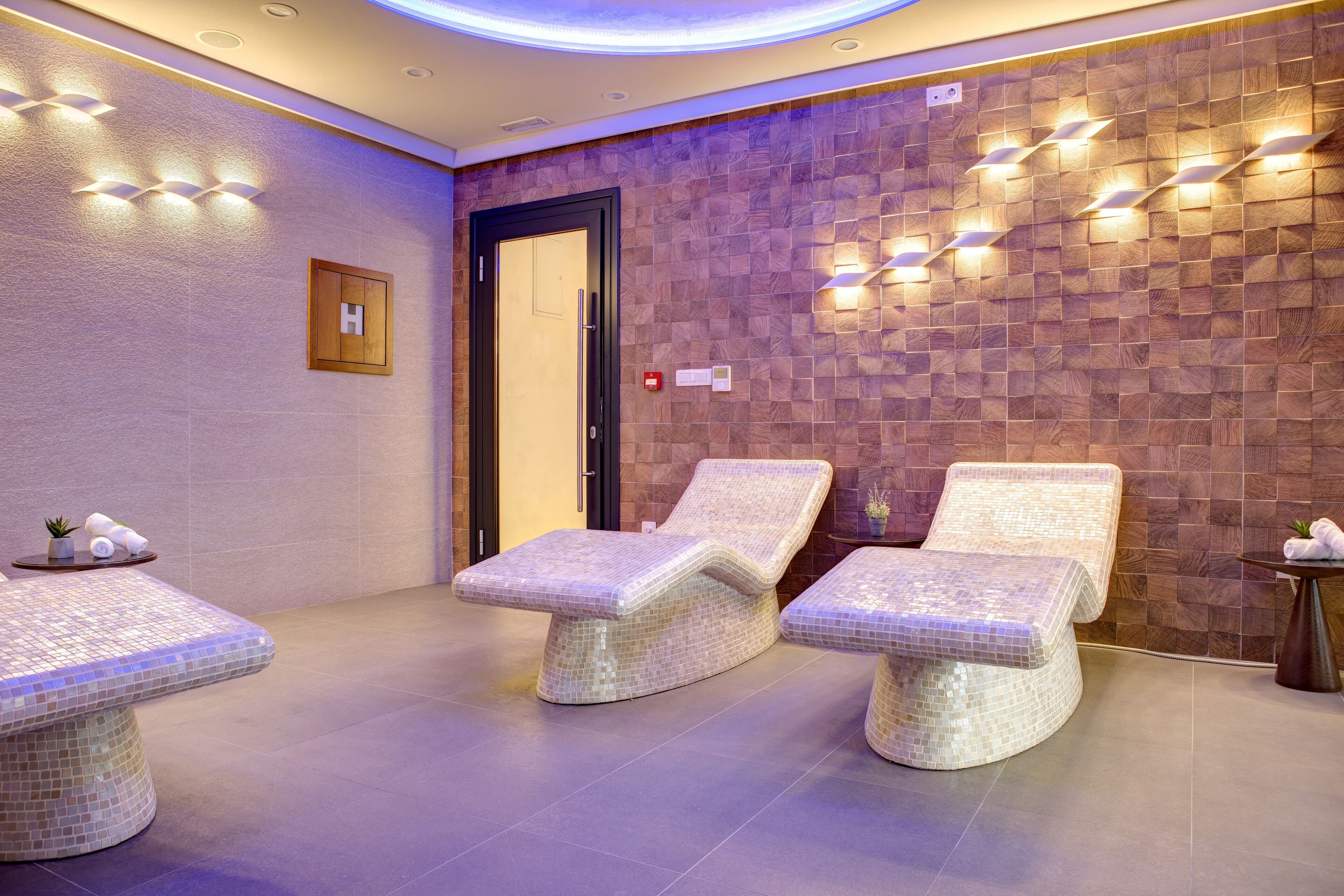 Um espaço de spa com três espreguiçadeiras revestidas em mosaico, parede de madeira com padrão herringbone e luminárias fixadas na parede.