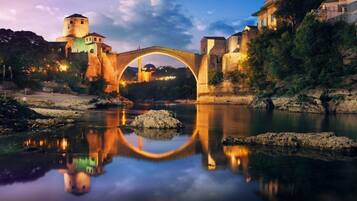 A stone bridge over a calm river with a reflection, surrounded by historic buildings and a tower.