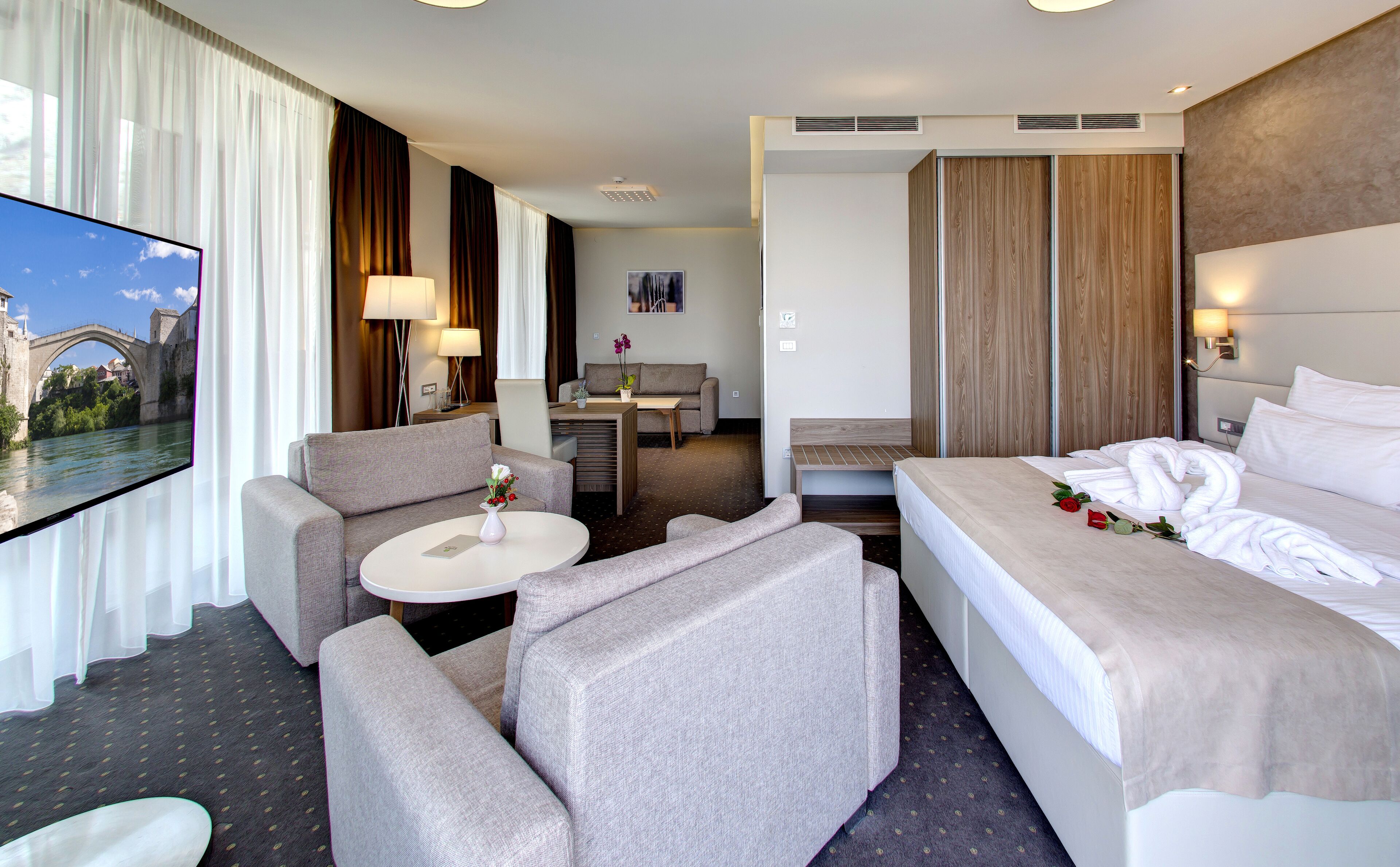 A hotel room with a bed, two armchairs, a small table, and a TV displaying a scenic view.