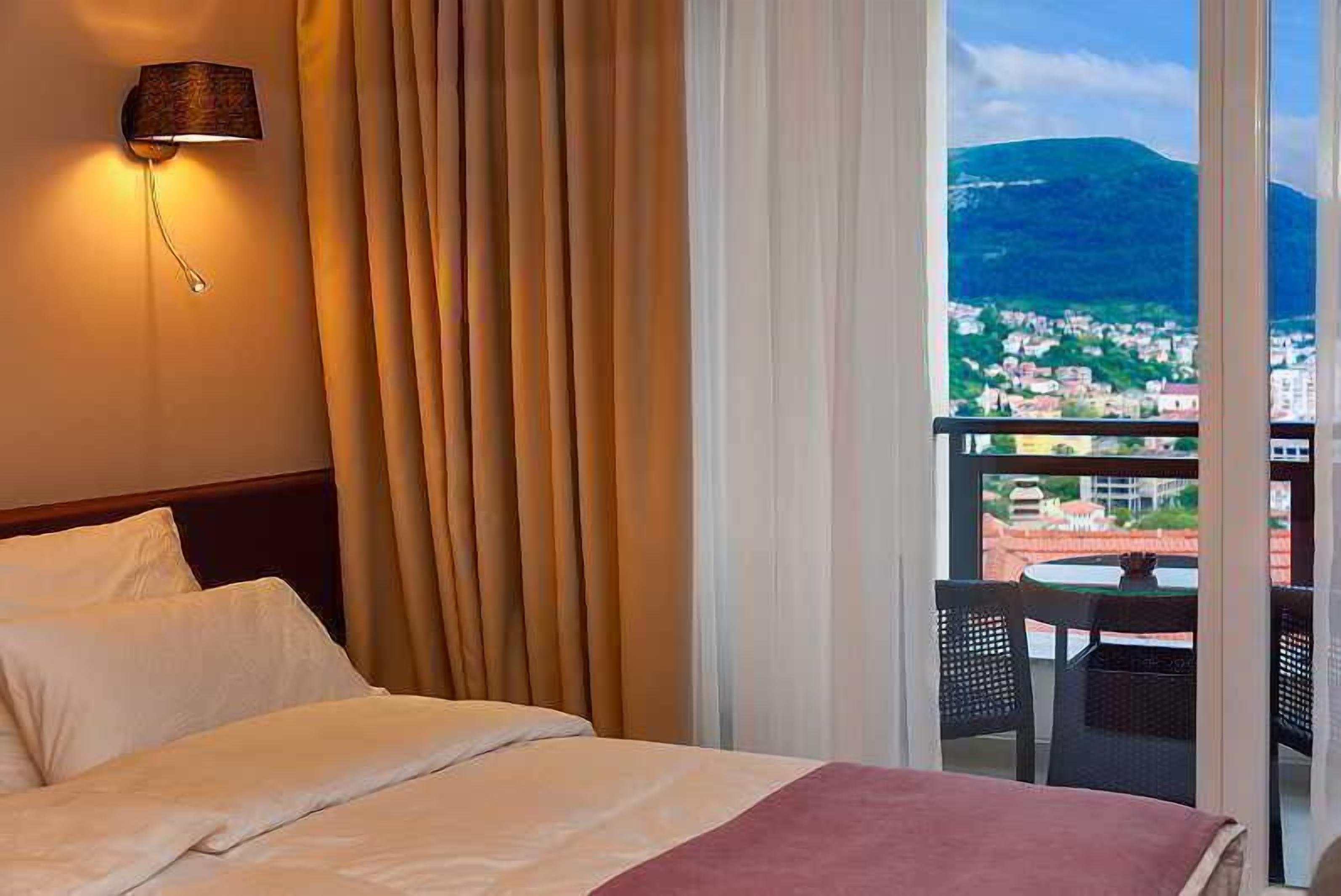 A hotel room with a bed, a wall-mounted lamp, a view of a town and mountains through large windows, and a balcony with a table and chairs.